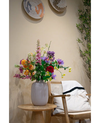 A white vase with Artificial Dogwood Real Touch Light Yellow 74cm stands on a wooden side table next to a light wooden chair with a white cushion and a rug with black edges, accentuated by woven wall decoration and a green plant in the background.