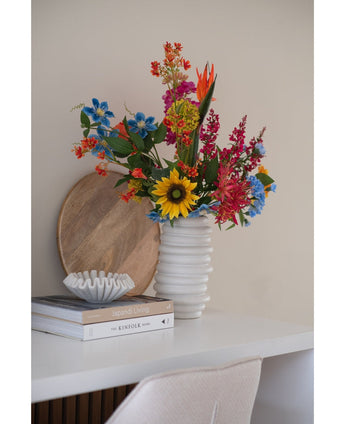 An Artificial Carnation flower bunch 70 cm blue in a white ceramic vase stands on a white desk next to stacked books and a small decorative bowl, with a round wooden tray leaning against the wall behind it.