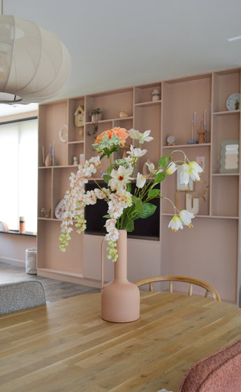A modern dining room with a wooden table with a peach-colored vase containing the Artificial flower Fritillary 94 cm white. In the background is a large built-in shelving unit decorated with stylish items.