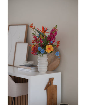 A white ribbed vase with Artificial Flower Catnip 75 cm pink stands on a white desk with books, framed art, a wooden tray and a cutting board, next to a beige chair in a modern minimalist room.