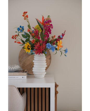 A white textured vase holds an Artificial Flower Catnip 75cm pink bouquet on a white table next to stacked books and decor, with a round wooden tray leaning against the beige wall in the background.