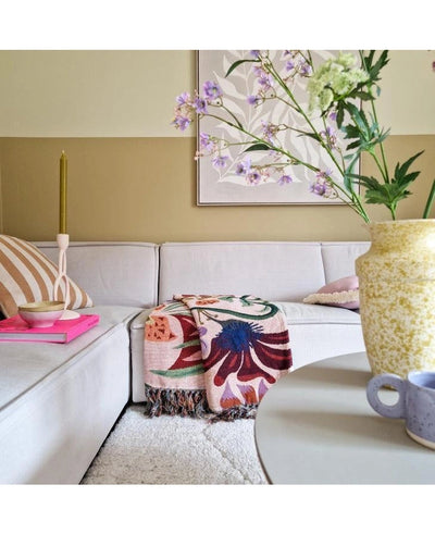 Een gezellige woonkamer met een lichtbeige bankstel, een gebloemde plaid, een roze kaars, een gestreept kussen en een Kunstbloem Kantbloem 76 cm wit in een gele vaas op de ronde tafel. Abstracte kunst siert de muur erachter.
