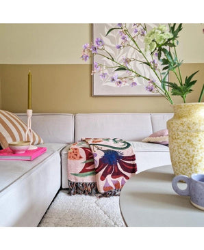 Een gezellige woonkamer met een lichtbeige bankstel, een gebloemde plaid, een roze kaars, een gestreept kussen en een Kunstbloem Kantbloem 76 cm wit in een gele vaas op de ronde tafel. Abstracte kunst siert de muur erachter.