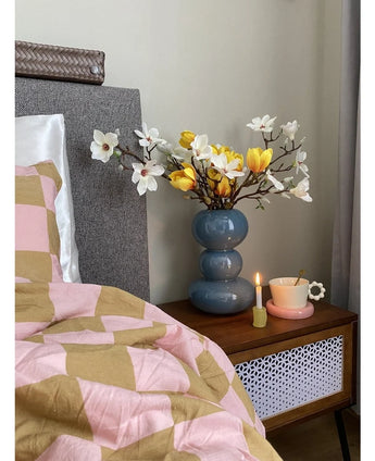 A bedside table with a blue vase containing the Artificial Flower Magnolia 50 cm yellow/orange, a pink teacup on a daisy-shaped saucer and a burning candle stands next to a bed covered with a pink and brown blanket with geometric patterns.