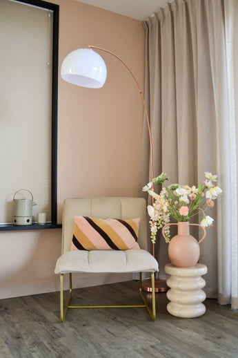 Een moderne, gezellige hoek met een crèmekleurige stoel met een gestreept kussen, een hoge boogvormige vloerlamp, beige gordijnen, een houten vloer en een ronde vaas met Kunstbloem Fritillary 94 cm wit bloemen op een gebeeldhouwde bijzettafel.