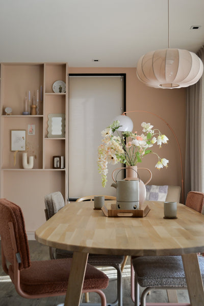 A modern dining room with a light wooden table, four chairs with cushions, the Artificial Flower Fritillary 94 cm white in a vase, ceramic cups, pastel pink walls, built-in shelves with decor and a large round hanging lamp above your head.