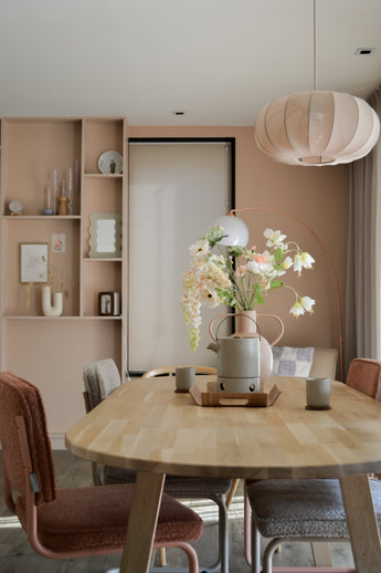 Een moderne eetkamer met een lichthouten tafel, vier stoelen met kussens, de Kunstbloem Fritillary 94 cm wit in een vaas, keramische kopjes, pastelroze muren, ingebouwde planken met decor en een grote ronde hanglamp boven je hoofd.