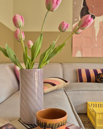 Een Kunstbloem Franse Tulp van 63 cm in roze staat in een vaas op de tafel naast een oranje kopje in een gezellige woonkamer met een crèmekleurige bank, kussens met patronen, een gele krat en abstracte wandkunst op de achtergrond.