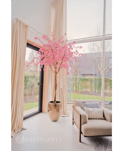 The Artificial Cherry Blossom Pink Tree 240 cm stands in a decorative pot near a large window in a bright, modern living room with cream-colored curtains, a beige armchair and a view of the garden outside.