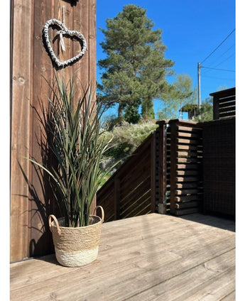 The Artificial Grass Bordeaux UV 120cm sits in a woven basket on a wooden deck near a wooden wall with a heart-shaped wreath, with trees and a clear blue sky in the background.