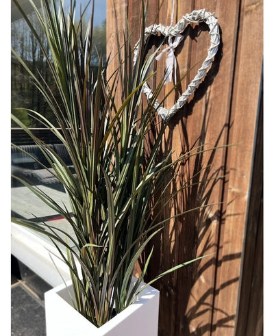 The Artificial Grass Bordeaux UV 120cm, a tall green artificial plant, stands next to a wooden wall above which hangs a white woven heart that casts sunlit shadows.