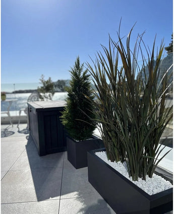 Artificial grass Bordeaux UV 120cm in rectangular planters with white stones decorates a modern outdoor terrace against a background of blue sky and trees in the distance.