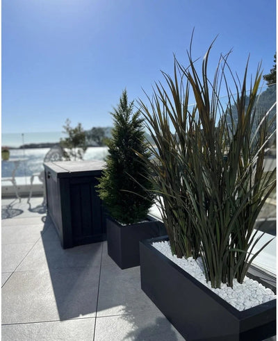 Artificial grass Bordeaux UV 120cm in rectangular planters with white stones decorates a modern outdoor terrace against a background of blue sky and trees in the distance.