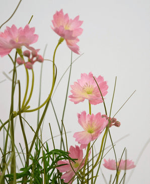 De Kunstgras roze Gesang Bloem uiengraspot 56cm brandvertragend heeft delicate roze bloemen met slanke groene stelen en smalle bladeren, gerangschikt tegen een lichte achtergrond voor een zacht, luchtig effect.