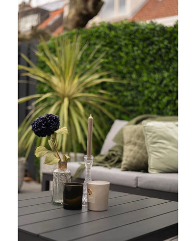 A modern outdoor table with a black surface shows a vase with a dark flower, a glass candle holder and two cups. Behind it is a sofa with green cushions opposite a lush green wall made of the Kunsthaag mat mix 50x50 cm.
