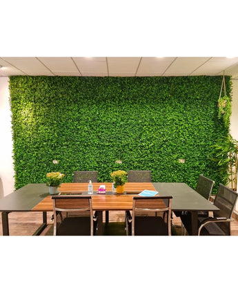 A modern meeting room features a long table, black chairs, small potted plants, notebooks and water bottles. The Kunsthaag mat mix 50x50 cm creates a lush background of green walls and gives the room a fresh and natural look.