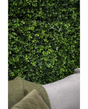 Close-up of a lush green artificial hedge mat mix 50x50 cm wall with vibrant leaves, with the corner of a light gray sofa and part of a green coat visible in the lower foreground.