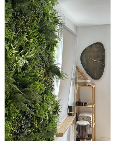 A lush Artificial Hedge Freshness 100x100 cm UV vertical garden covers one wall of a bright room. Nearby, a modern triangular wall clock hangs above a wooden shelf with towels, a djembe drum, baskets and small decorative pieces.