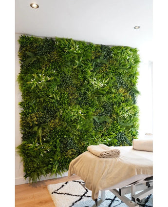A massage table with folded towels stands in front of a Kunsthaag Freshness 100x100 cm UV vertical garden in a bright, modern room with white walls and a geometric carpet.