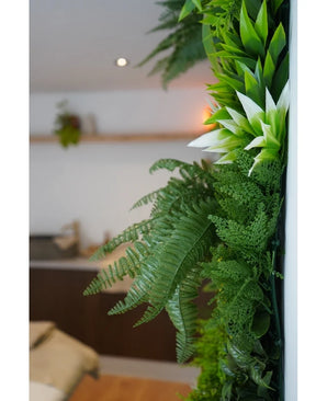 A close-up of lush green Artificial Hedge Freshness 100x100 cm UV artificial plants adorns a vertical interior wall, with a blurred background showing neutral furniture and a small hanging plant.