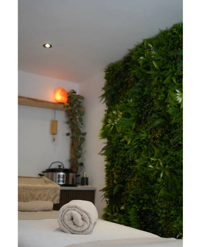 A tranquil spa area with a massage table, soothing lighting, a Himalayan salt lamp on a shelf and a lush Artificial Hedge Freshness 100x100 cm UV green plant wall for extra serenity.