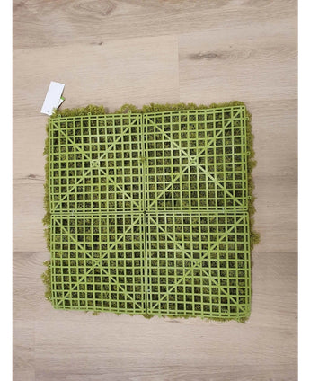 De Kunsthaag Nordic Mos 50x50 cm UV is een groene kunstgrasmat met een zichtbare kunststof rasterrug, ideaal voor luxe interieurs of het creëren van een groene muur. Hij rust op een lichte houten vloer met een klein wit label in een hoek.