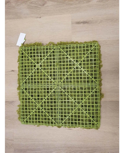 De Kunsthaag Nordic Mos 50x50 cm UV is een groene kunstgrasmat met een zichtbare kunststof rasterrug, ideaal voor luxe interieurs of het creëren van een groene muur. Hij rust op een lichte houten vloer met een klein wit label in een hoek.