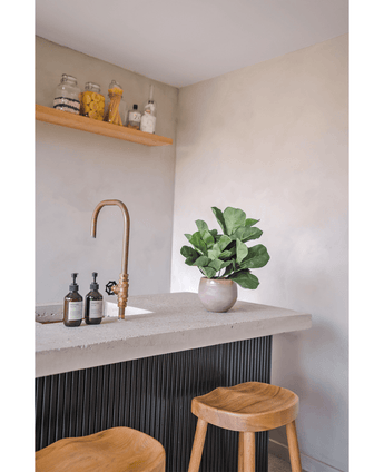 Een modern aanrecht met een messing kraan, twee zeepdispensers, de Kunstplant Tabaksplant Fiddle Leaf UV 45 cm potplant en twee houten krukjes. Een zwevende plank met potten en flessen is gemonteerd op een lichtgekleurde muur.