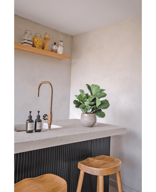 A modern countertop with a brass tap, two soap dispensers, the Artificial Tobacco Plant Fiddle Leaf UV 45 cm potted plant and two wooden stools. A floating shelf with jars and bottles is mounted on a light-colored wall.