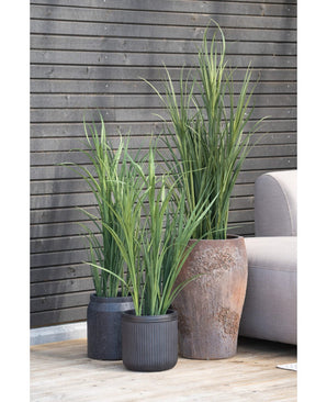 Drie Kunstgras Groen UV 150cm kunstplanten met lange, smalle bladeren staan op een houten vlonder naast een lichtgrijze buitenbank, tegen een donkere houten lattenwand.