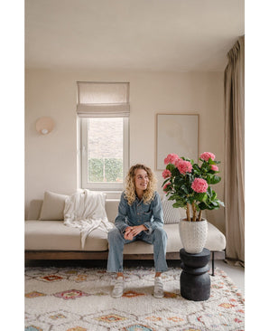 Een vrouw in spijkerpak zit op een beige bank in een lichte woonkamer. Op de voorgrond staat de Kunstplant Hortensia UV 85 cm roze, een grote roze kunsthortensia in pot op een zwarte standaard. Zonlicht stroomt door het raam achter haar.