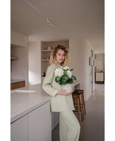 A woman with curly blonde hair, dressed in a light green outfit, stands in a modern kitchen with an Artificial Hydrangea White UV 35 cm plant in her hands – with creamy white flowers – and smiles at the camera.