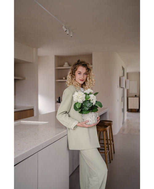 A woman with curly blonde hair, dressed in a light green outfit, stands in a modern kitchen with an Artificial Hydrangea White UV 35 cm plant in her hands – with creamy white flowers – and smiles at the camera.