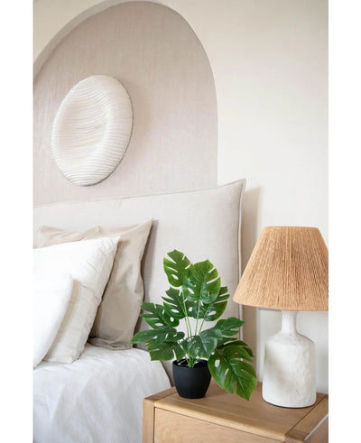 A modern bedroom in neutral tones has a white bed with beige and white pillows, a wooden bedside table with a ceramic lamp and a woven shade, abstract wall art above the bed and the Art Monstera 40 cm in a black decorative pot that adds greenery.