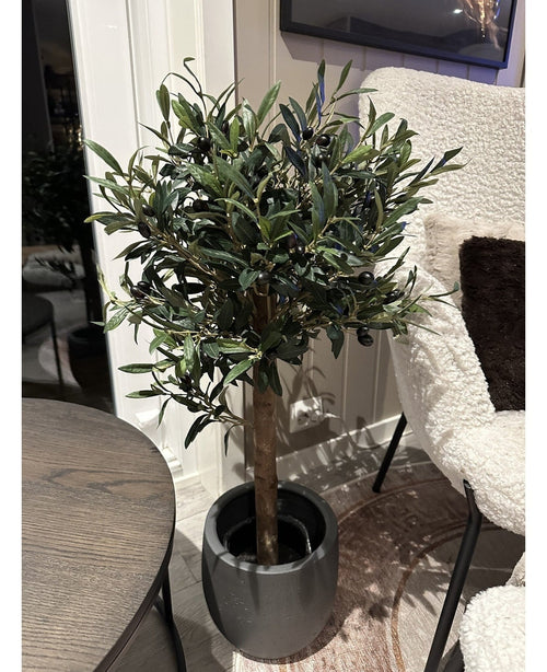 The Artificial olive tree UV 85 cm, a compact and easy-care artificial plant with dark green leaves and black olives, stands indoors next to a round wooden table and an armchair with a white texture.