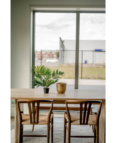 Een houten tafel met twee stoelen, een Kunstpalm Parlour UV 60 cm in een pot en een gouden vaas staan bij een groot raam met uitzicht op een industriegebied met gebouwen en een hek.