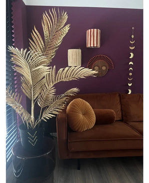 A modern living room with a brown velvet sofa, round folding cushion and the Areca gold artificial palm 120 cm in a black vase as a gold decor. Geometric wall art hangs on a dark purple wall, with wooden floors and vertical slats in view.
