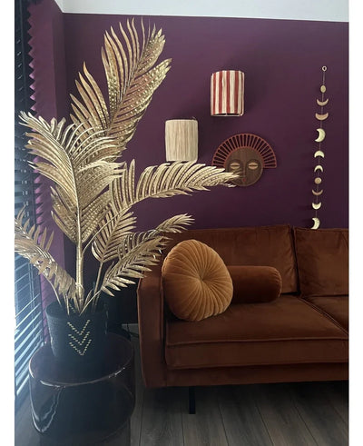 A modern living room with a brown velvet sofa, round folding cushion and the Areca gold artificial palm 120 cm in a black vase as a gold decor. Geometric wall art hangs on a dark purple wall, with wooden floors and vertical slats in view.