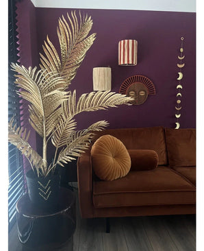Een moderne woonkamer met een bruin fluwelen bank, rond plooikussen en de Kunstpalm Areca goud 120 cm in een zwarte vaas als gouddecor. Geometrische wandkunst hangt op een donkerpaarse muur, met houten vloeren en verticale lamellen in het zicht.
