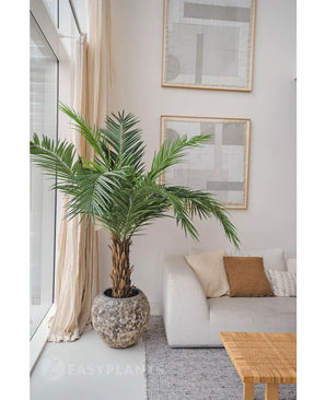 De Kunstpalm Phoenix Canarie 160 cm voegt een tropische sfeer toe naast een lichte bank met neutrale kussens in een moderne woonkamer met abstracte kunst, transparante gordijnen en een geweven salontafel.