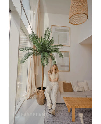 A woman smiles with a drink in a bright, modern living room, standing next to the Artificial Palm Phoenix De Luxe 210 cm. The space features large windows, neutral décor, framed art, a sofa, a wooden table and woven accents.