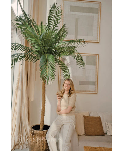 A woman with blond hair wearing a beige top and light trousers smiles as she leans against the Artificial Palm Phoenix De Luxe 210 cm in a bright, modern living room with a neutral decor and abstract art on the walls.