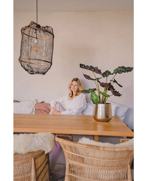 Een vrouw in een witte blouse zit aan een houten eettafel met rieten stoelen. De gezellige, zacht verlichte kamer heeft de Kunstplant Alocasia UV 67 cm en een hanglamp met textuur als decor.