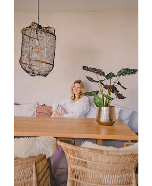 A woman in a white blouse sits at a wooden dining table with wicker chairs. The cozy, softly lit room has the Artificial Plant Alocasia UV 67 cm and a textured hanging lamp as decor.
