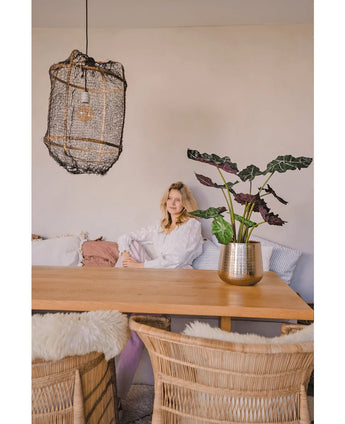 A woman in a white blouse sits at a wooden dining table with wicker chairs. The cozy, softly lit room has the Artificial Plant Alocasia UV 67 cm and a textured hanging lamp as decor.