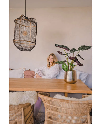 A woman in a white blouse sits at a wooden dining table with wicker chairs. The cozy, softly lit room has the Artificial Plant Alocasia UV 67 cm and a textured hanging lamp as decor.