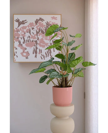 The Artificial Apple Leaf 90 cm, a green-leaved pot plant in a pink ribbed pot, stands on a beige plinth in front of a wall with abstract pink floral art and a translucent white curtain on the right.