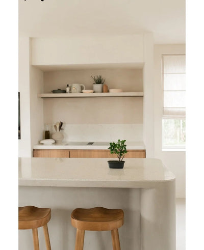 A minimalist kitchen with light walls and cupboards, open shelves with plates and a plant, two wooden stools near a curved island and the Artificial Bonsai Ficus 30 cm plant on the counter near a bright window.