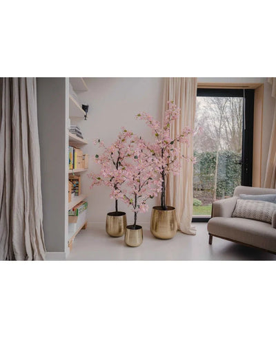 Een Kunstplant Bougainvillea 120 cm roze staat bij het raam in een gezellige woonkamer met witte muren, een boekenplank, beige gordijnen en een grijze fauteuil.