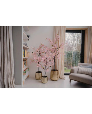 Een Kunstplant Bougainvillea 120 cm roze staat bij het raam in een gezellige woonkamer met witte muren, een boekenplank, beige gordijnen en een grijze fauteuil.